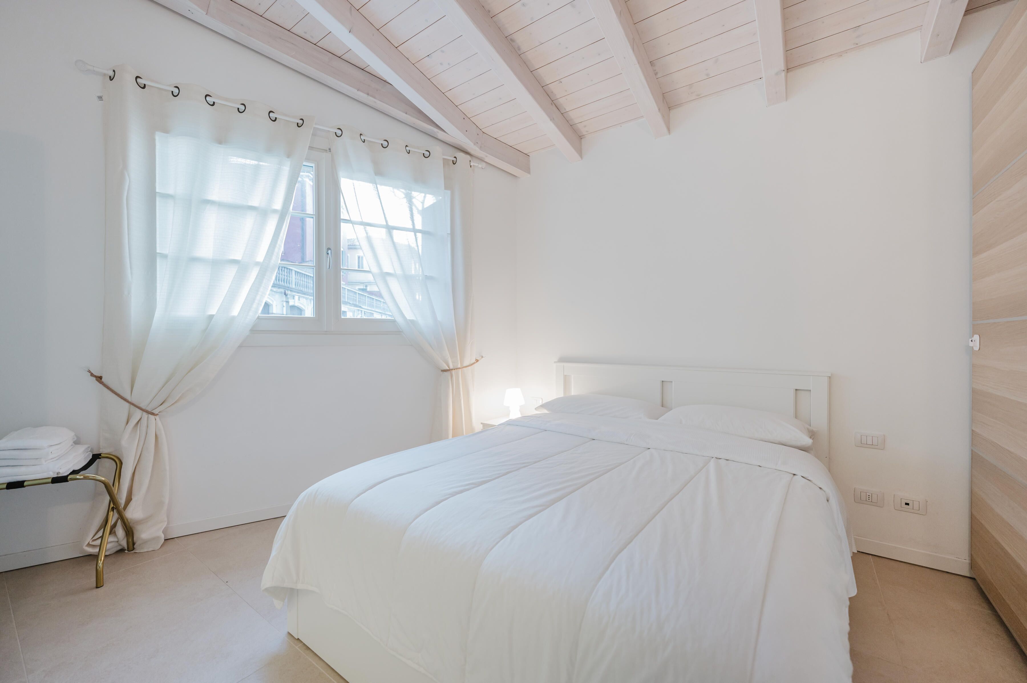A bedroom with a bed, a window with sheer curtains, a bedside table, and a lamp.