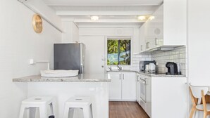 Eine kleine Küche mit weißen Schränken, einem Edelstahlkühlschrank, einer Kaffeemaschine und einem Fenster mit Blick auf Grünflächen.