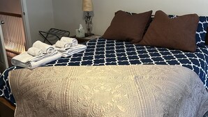 Ein ordentlich bezogenes Bett mit einem gemusterten Überwurf und Kissen, eine Nachttischlampe und ein gefaltetes Handtuch auf dem Nachttisch.