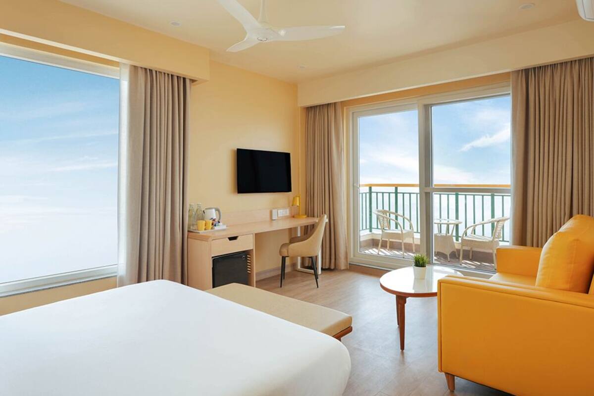 Una habitación de hotel con cama, escritorio, silla, sofá amarillo, televisor y balcón con vistas.
