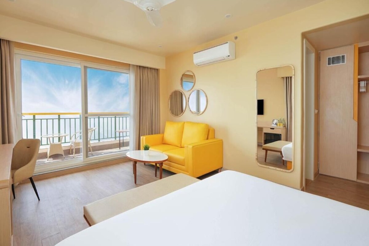 A hotel room with a bed, a yellow sofa, a small table, a balcony with a view, and an air conditioning unit.