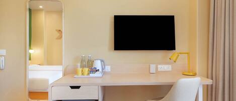Quarto de hotel com cama, escrivaninha com cadeira, televisão, mini-geladeira e espelho.