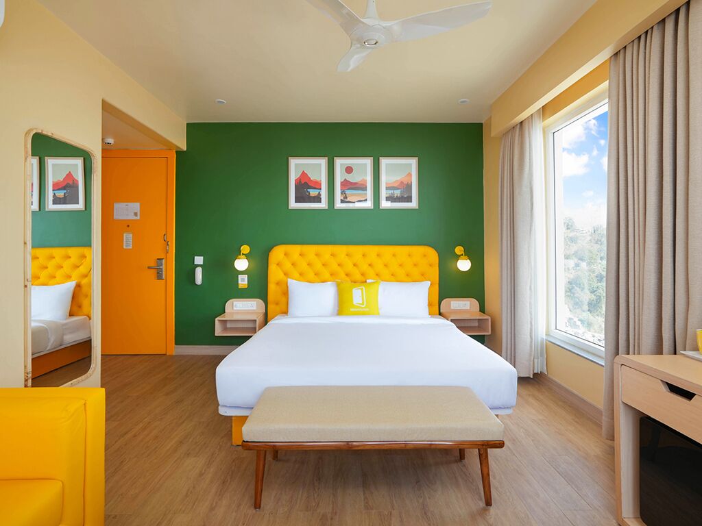 Habitación de hotel con cama, mesitas de noche, un banco, ventilador de techo, obras de arte en la pared y una ventana con cortinas.