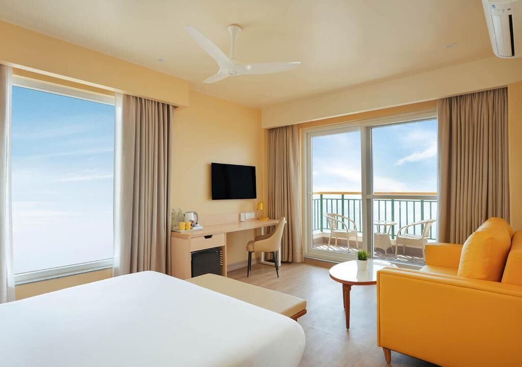 Habitación de hotel con cama, escritorio, silla, sofá amarillo, televisor y balcón con vistas.