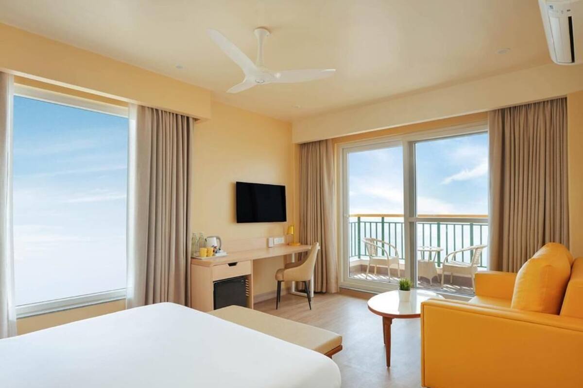 Habitación de hotel con cama, escritorio, silla, sofá amarillo, televisor y balcón con vistas.