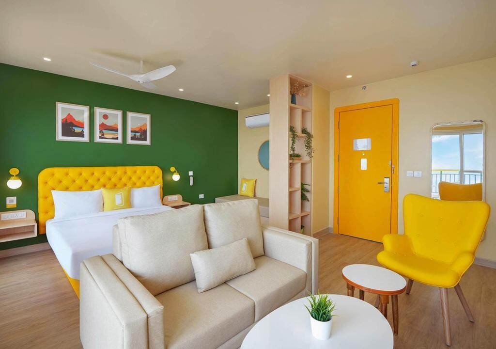 Habitación de hotel con cama, sofá, sillón, una mesita y pared verde decorada con cuadros.