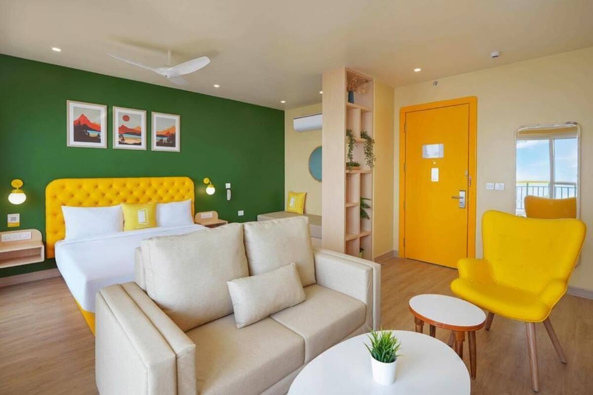 Habitación de hotel con cama, sofá, sillón, una mesita y pared verde decorada con cuadros.
