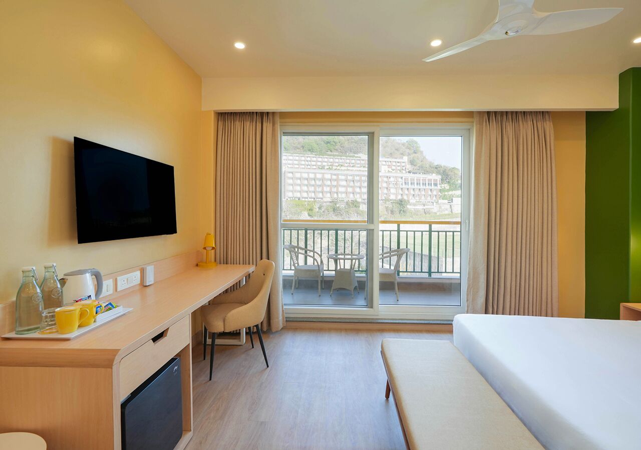 Habitación de hotel con cama, escritorio con silla, televisión y balcón con vista a edificios.