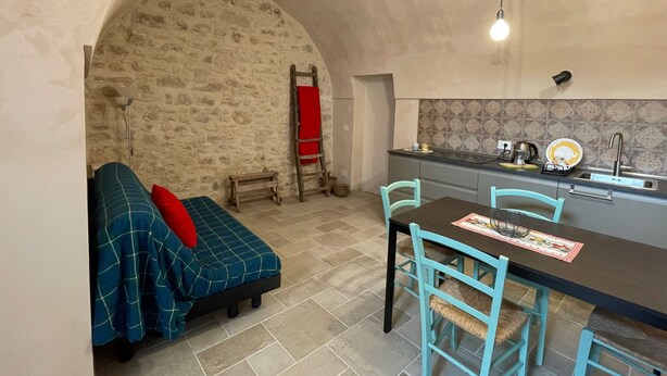 Ein gemütliches Zimmer mit Steinwand, einem Bett mit kariertem Überwurf, einem kleinen Tisch mit Stühlen und einer Küchenzeile mit Fliesenrückwand.
