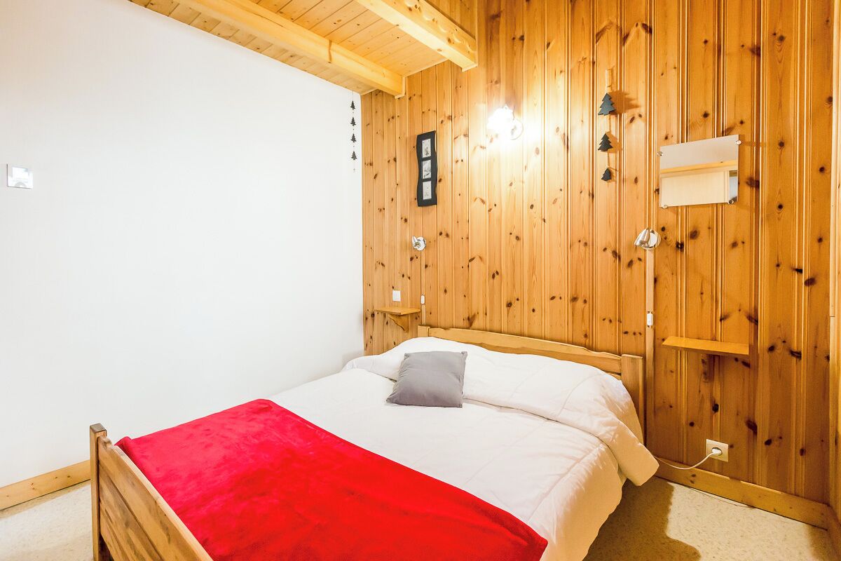 A wooden-paneled bedroom with a bed, a red blanket, a nightstand, and a wall-mounted shelf.