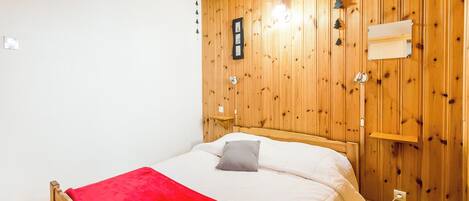 A wooden-paneled bedroom with a bed, a red blanket, a nightstand, and a wall-mounted shelf.