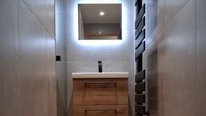 Un baño estrecho con tocador de madera, espejo con iluminación integrada y suelo de baldosas.