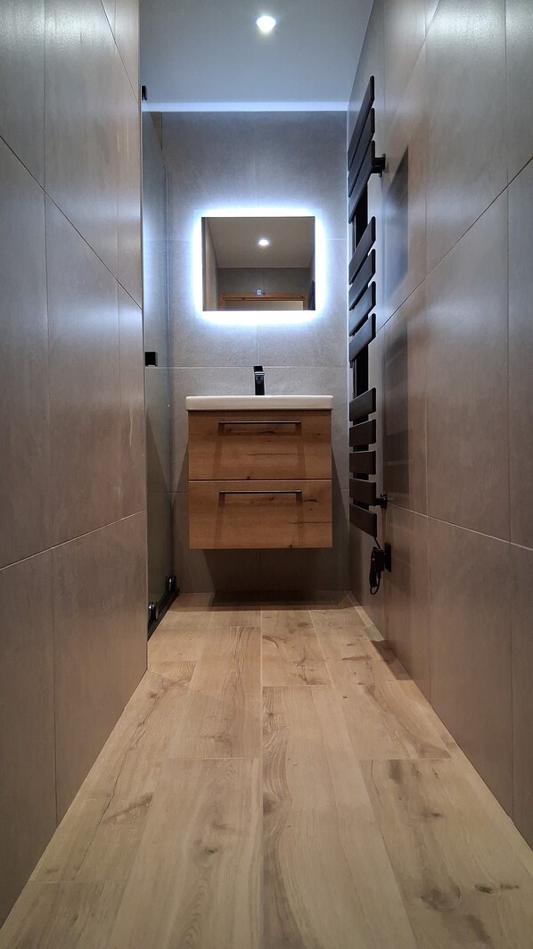 Une petite salle de bain avec un meuble-lavabo en bois, un miroir doté d’un éclairage intégré et un sol carrelé.