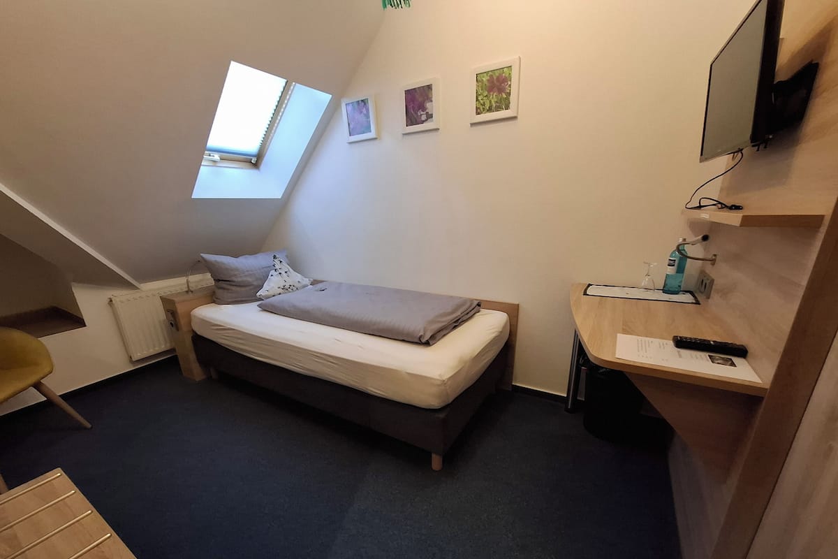 A small, compact room with a bed, a desk, a chair, a radiator, a window, and a TV mounted on the wall.