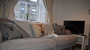 A grey sofa with patterned cushions, a flat-screen TV on a white stand, and a window with curtains.