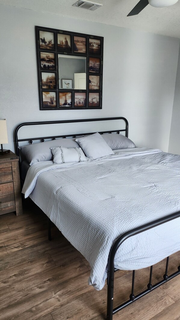 Een netjes opgemaakt bed met een gewatteerde deken, een nachtkastje met een lamp en een aan de muur bevestigd fotolijstje met foto's van een stadsgezicht.