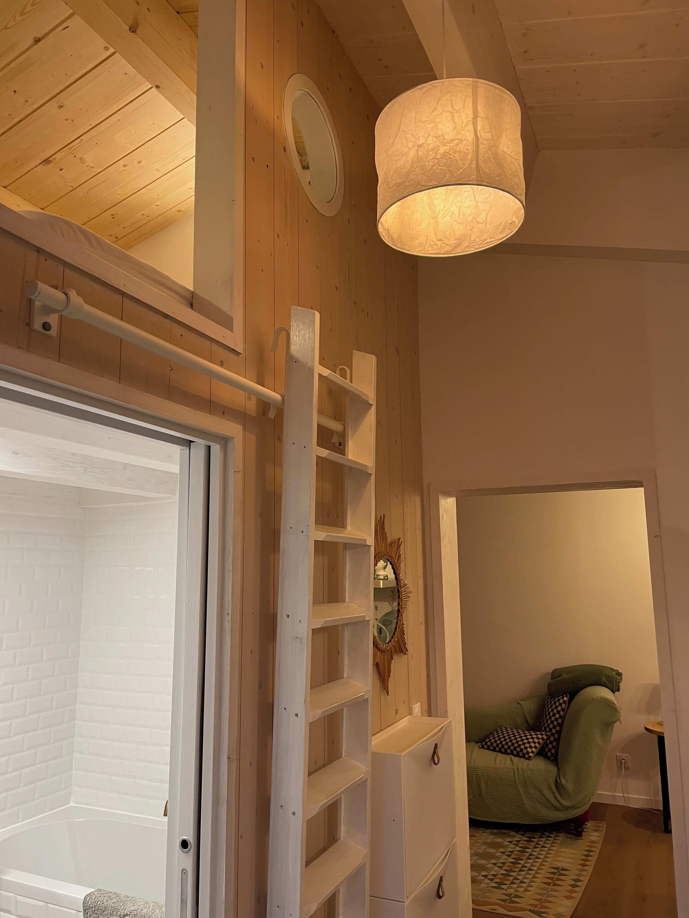 Une pièce avec un plafond en bois, une porte blanche, un mur blanc orné d’un miroir, une étagère blanche, un canapé vert et un tapis à motifs.