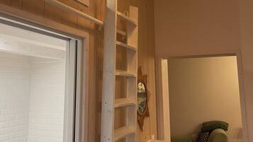 Une pièce avec un plafond en bois, une porte blanche, un mur blanc orné d’un miroir, une étagère blanche, un canapé vert et un tapis à motifs.