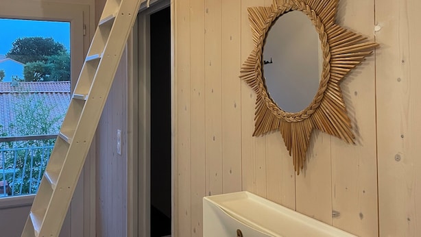 A bathroom with a wooden wall, a white sink, a mirror with a sunburst frame, and a ladder leading to an upper level. - Cosy, comfortable 4-star apartment (Vaux-sur-Mer)
