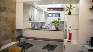 Une cuisine moderne avec des meubles blancs, un plan de travail noir et une étagère en verre où sont disposées des plantes. On y trouve deux tabourets de bar noirs et une chaise blanche.