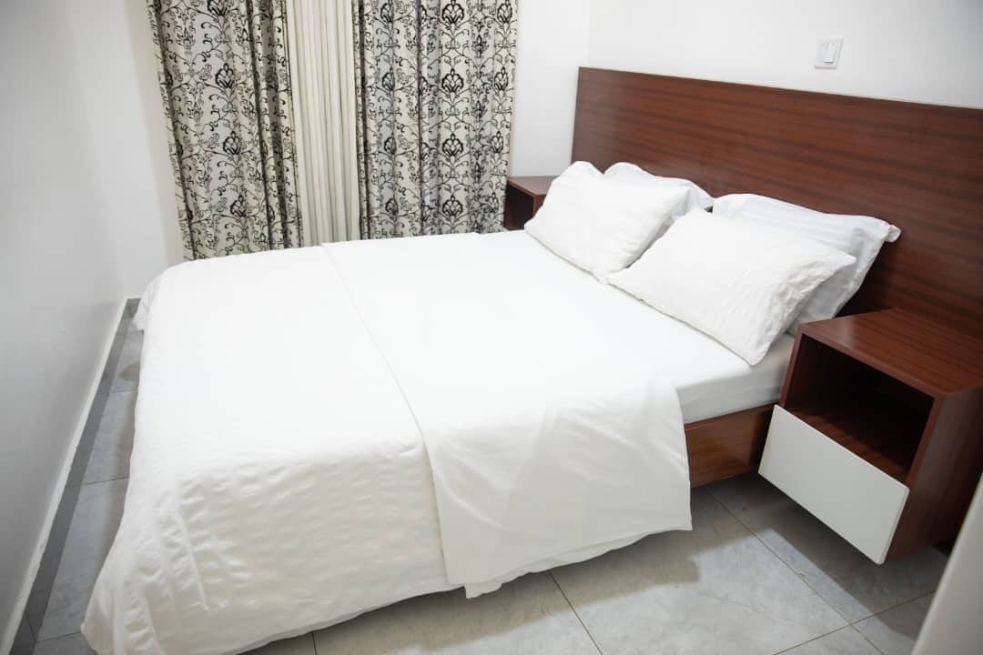A neatly made bed with white bedding and a wooden headboard. There is a wooden nightstand with a drawer on the right side of the bed.