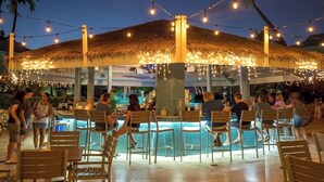 An outdoor bar area with a pool, surrounded by wooden chairs and tables, under a thatched roof with string lights. - Stunning 2 bedroom condo at Sunset Cove - Seven Mile beach (George Town)
