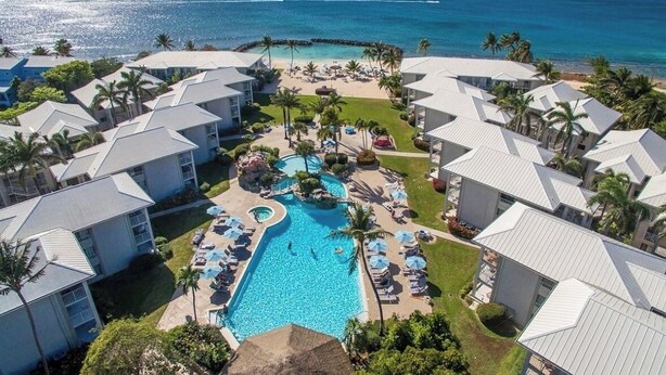 A resort with a central swimming pool surrounded by lounge areas, palm trees, and ocean views. - Stunning 2 bedroom condo at Sunset Cove - Seven Mile beach (George Town)