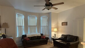 A living room with a ceiling fan, two windows with curtains, a red armchair, a black leather sofa, and a small table with a lamp. - Charming 4-bedroom family friendly house in Monongahela. Make yourself at home! (Monongahela)