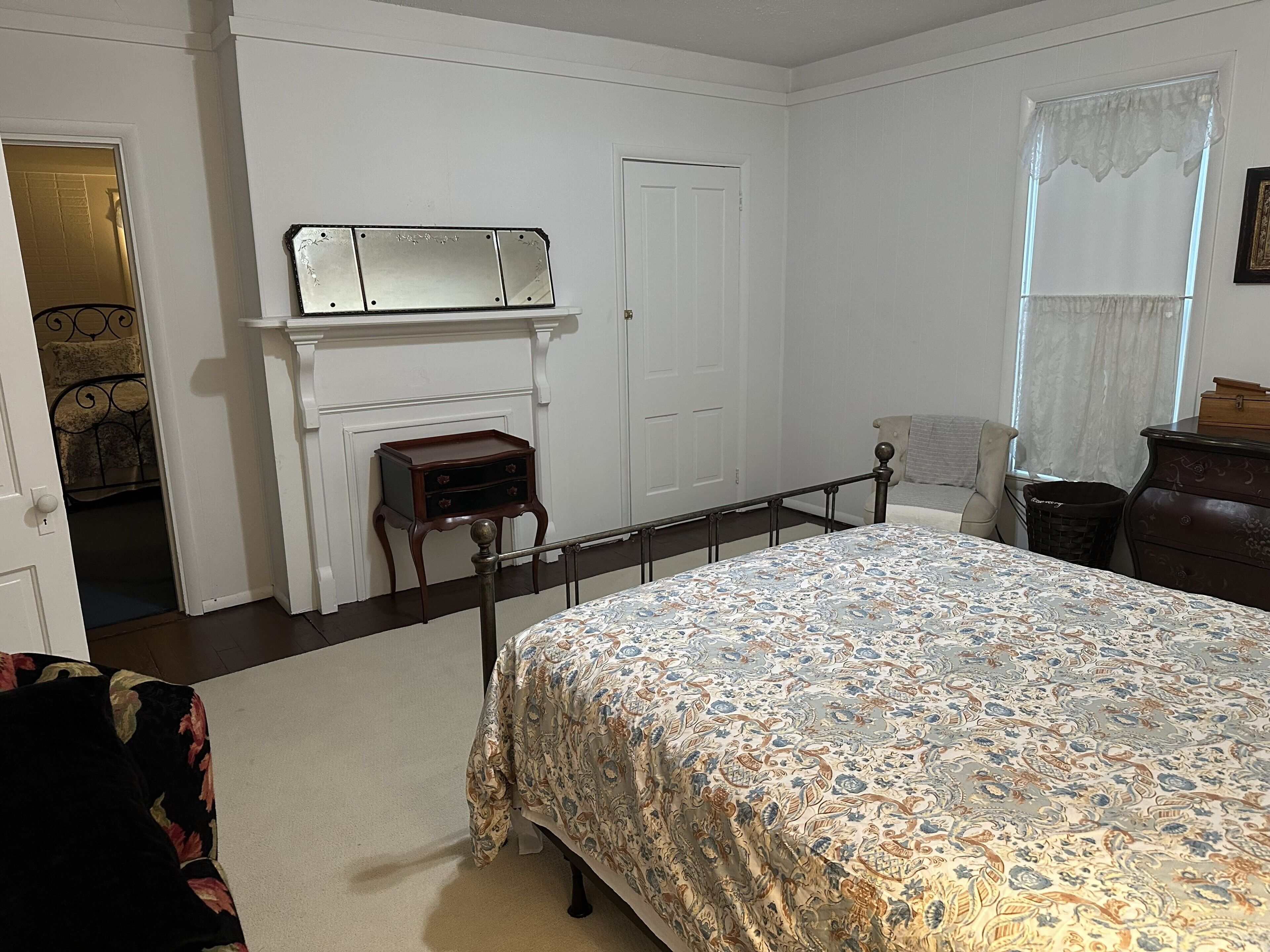 Une chambre à coucher comprenant un lit, une commode, une table de chevet, un miroir, une cheminée et une porte donnant sur une autre pièce.