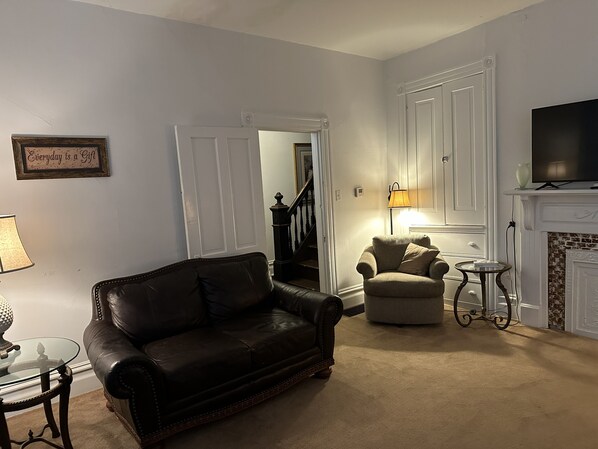 Un salon avec un canapé en cuir, une table basse en verre, un petit pouf, une lampe, un téléviseur et une cheminée.