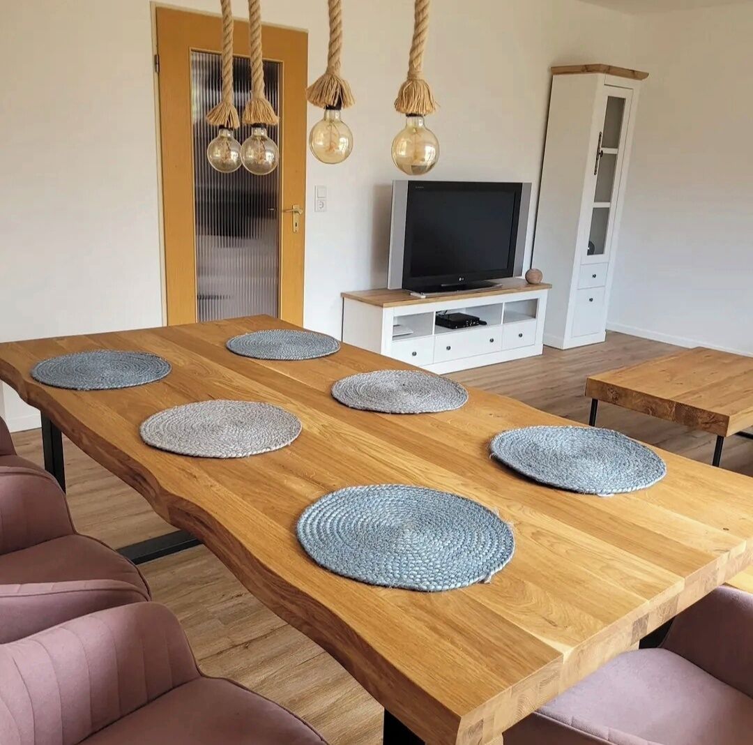 A dining area with a long wooden table, six grey placemats, a television on a white cabinet, and a hanging light fixture.