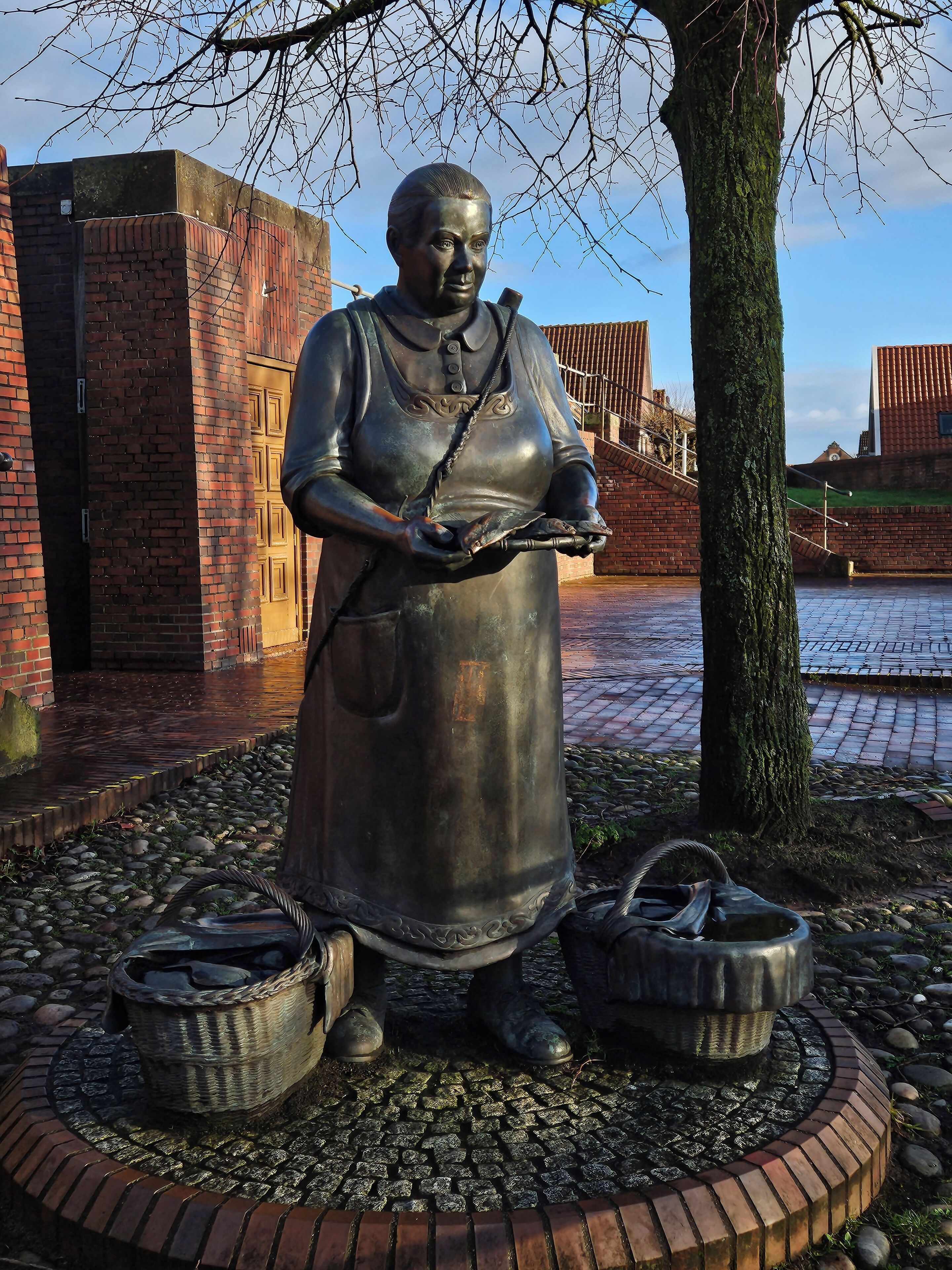 Een standbeeld van een persoon met een mand en een pot, omringd door een bakstenen rand en een boom.