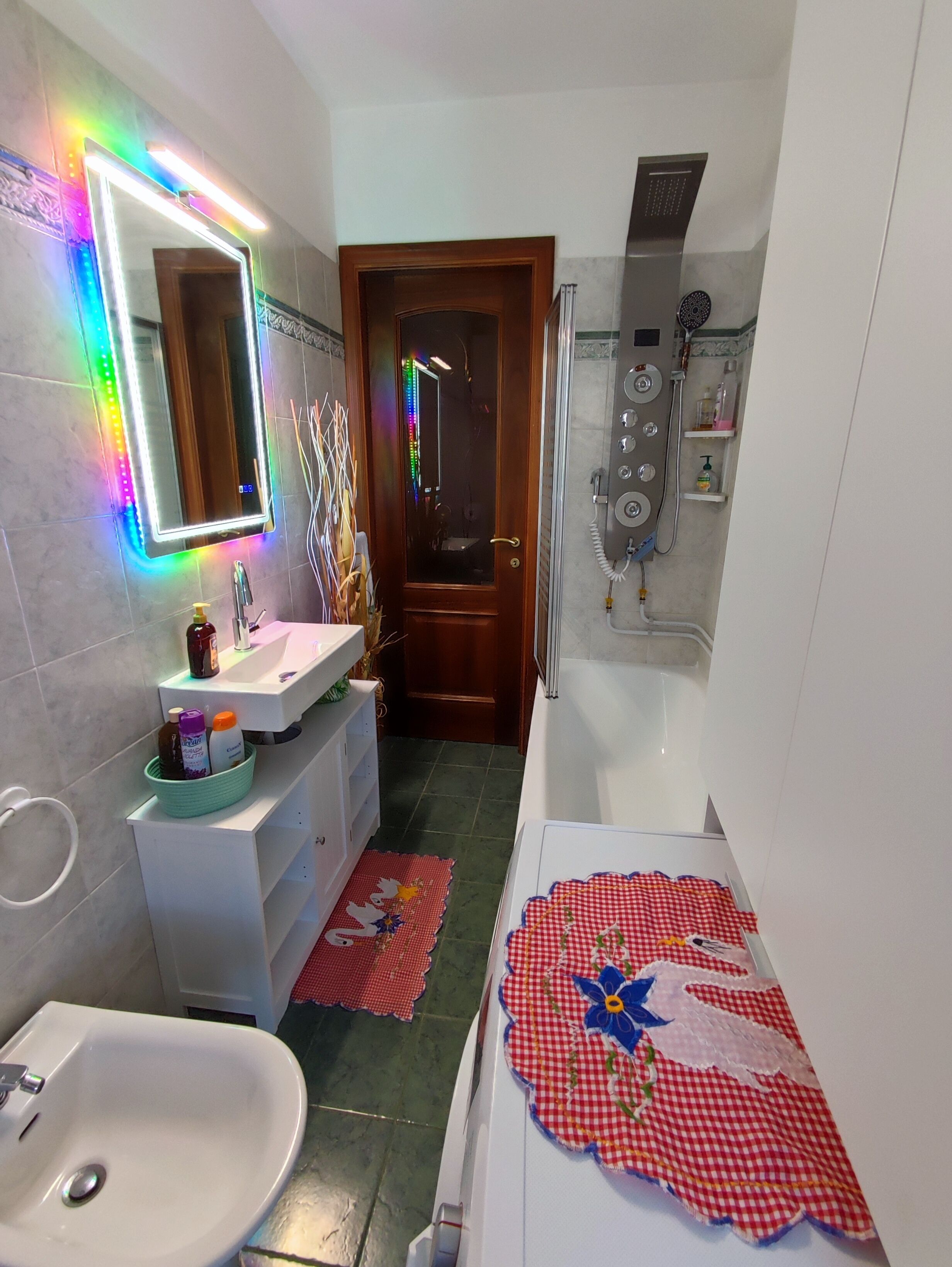 A bathroom with a white sink, a mirror with LED lights, a shower with a control panel, and a red checkered mat on the floor.
