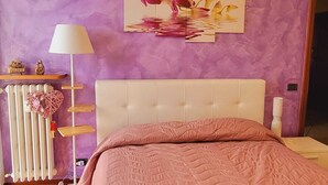 Un dormitorio con cabecera blanca, colcha acolchada rosa, radiador, lámpara y estante con adornos.