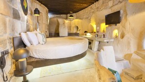 A circular bed in a stone-walled room with a stone bench, a small table, and a mounted TV. - EKA CAVE MAISON (Ürgüp)