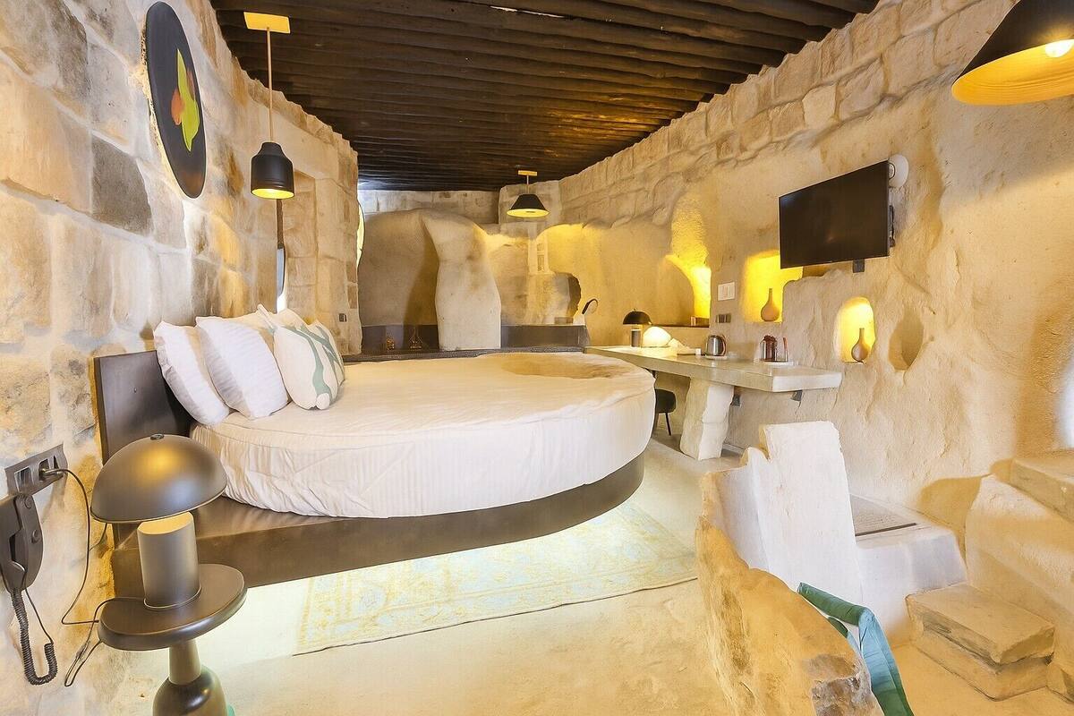 Ein rundes Bett in einem Raum mit Steinwänden, eine Steinsitzbank, ein kleiner Tisch und ein an der Wand befestigtes Fernsehgerät.