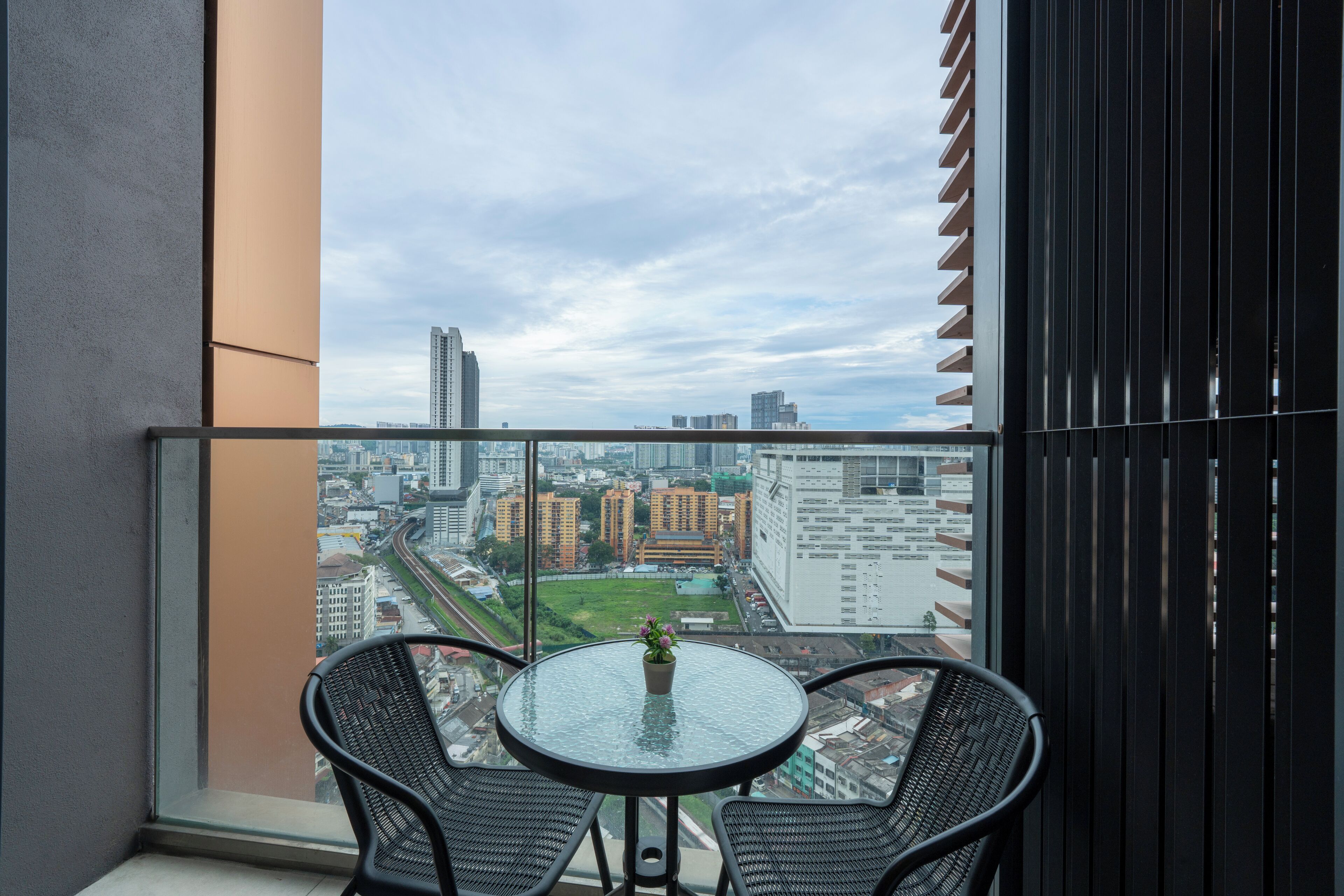 A balcony with a glass table and two chairs, overlooking a cityscape with buildings and a green park.