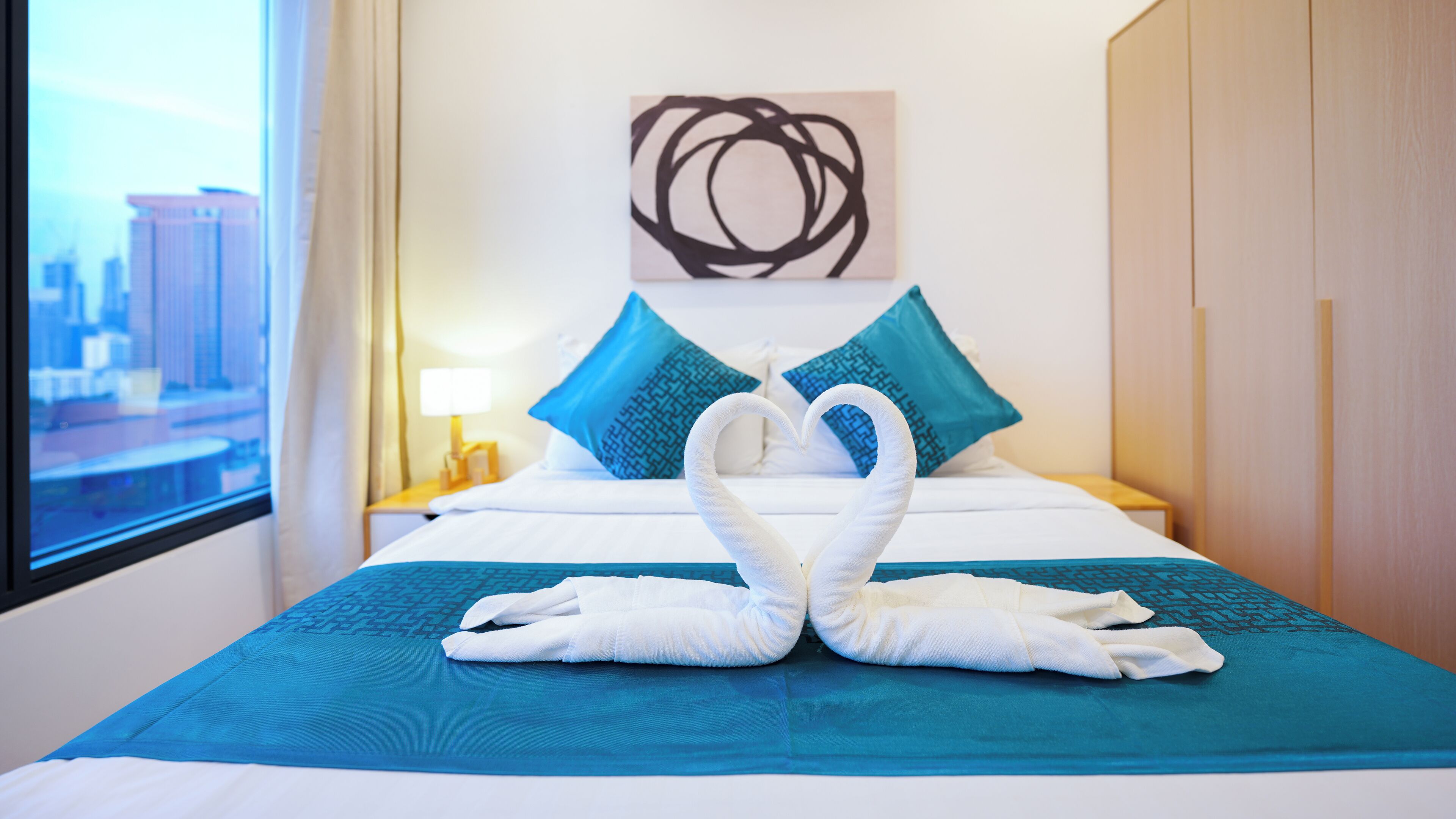 Hotelkamer met een bed, een raam met uitzicht op de stad, een kunstwerk aan de muur en een handdoekopstelling op het bed.