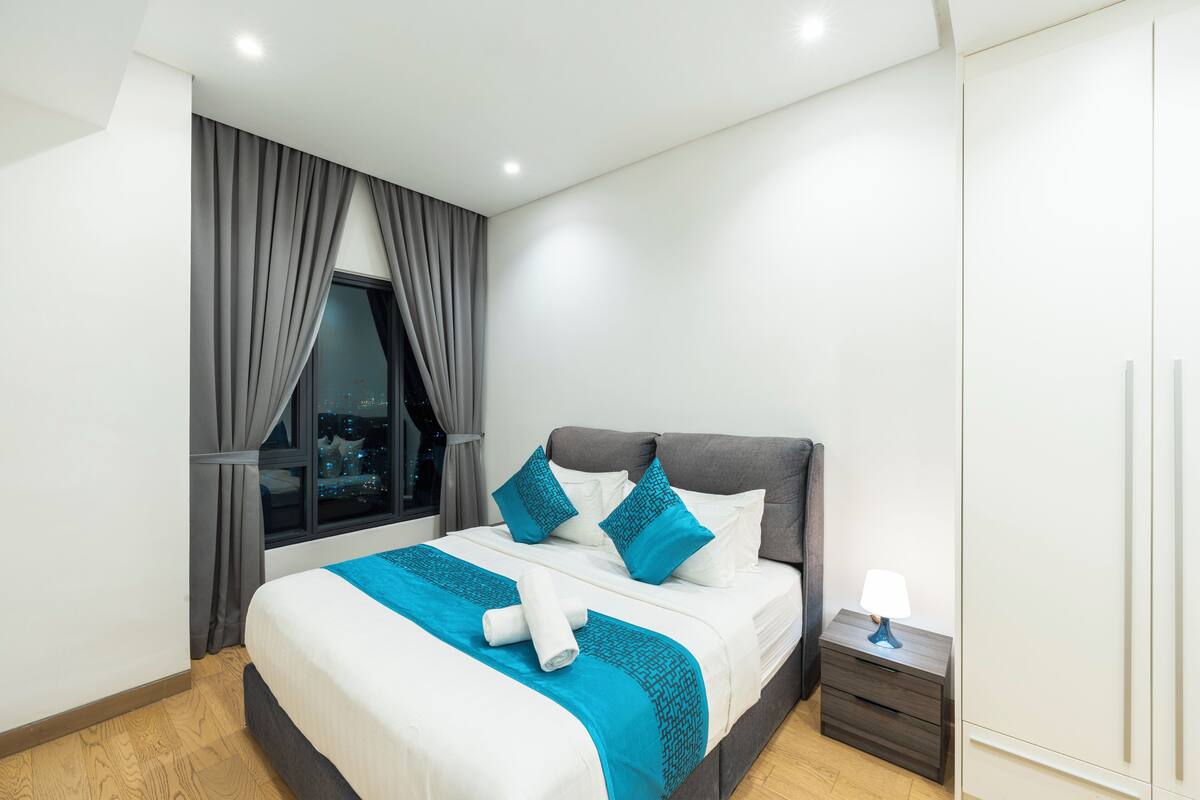 A modern hotel room with a large bed, grey curtains, a nightstand with a lamp, and a view of the city at night.