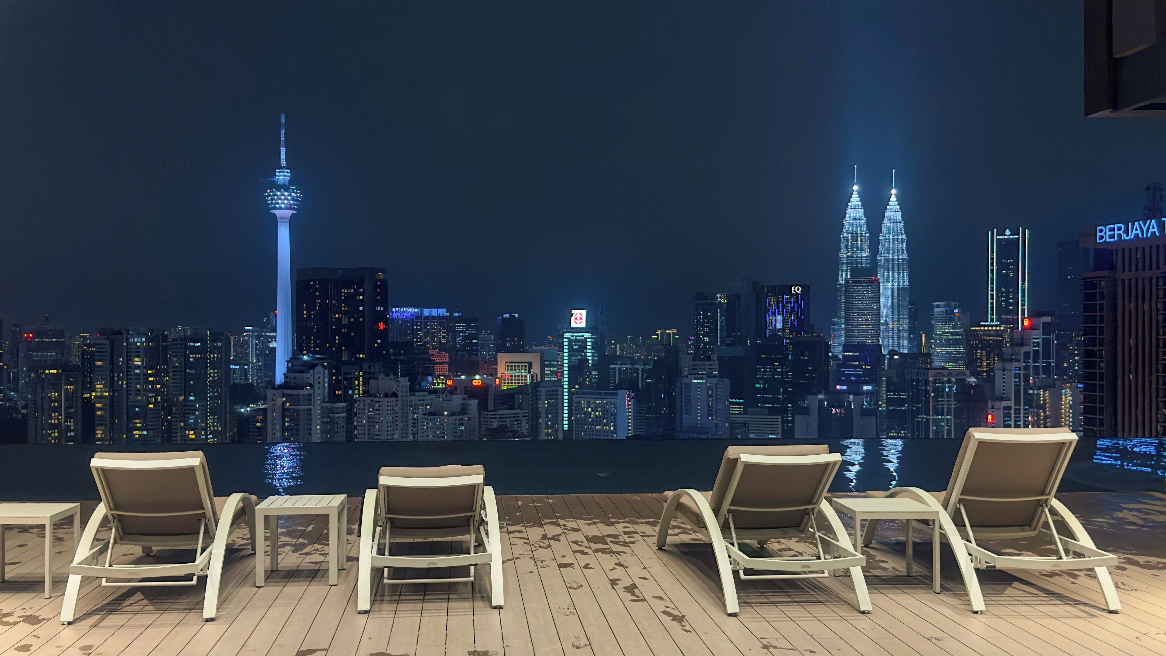 A rooftop lounge with four lounge chairs and a table, overlooking a city skyline at night.