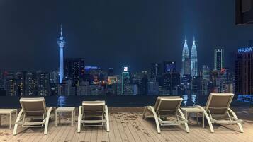 A rooftop lounge with four lounge chairs and a table, overlooking a city skyline at night.