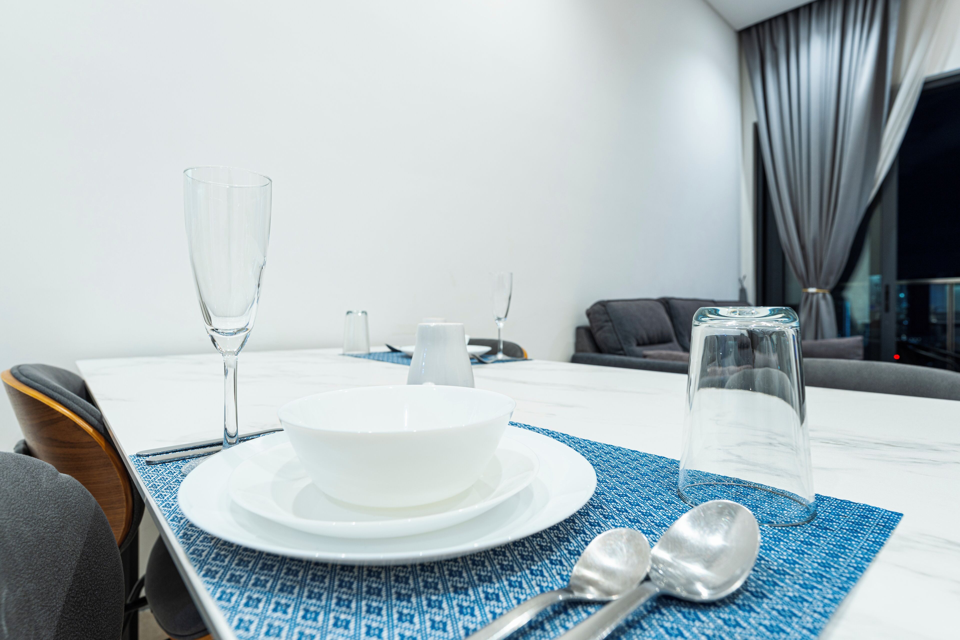 A dining table with a white bowl, two wine glasses, and a water glass on a blue placemat.