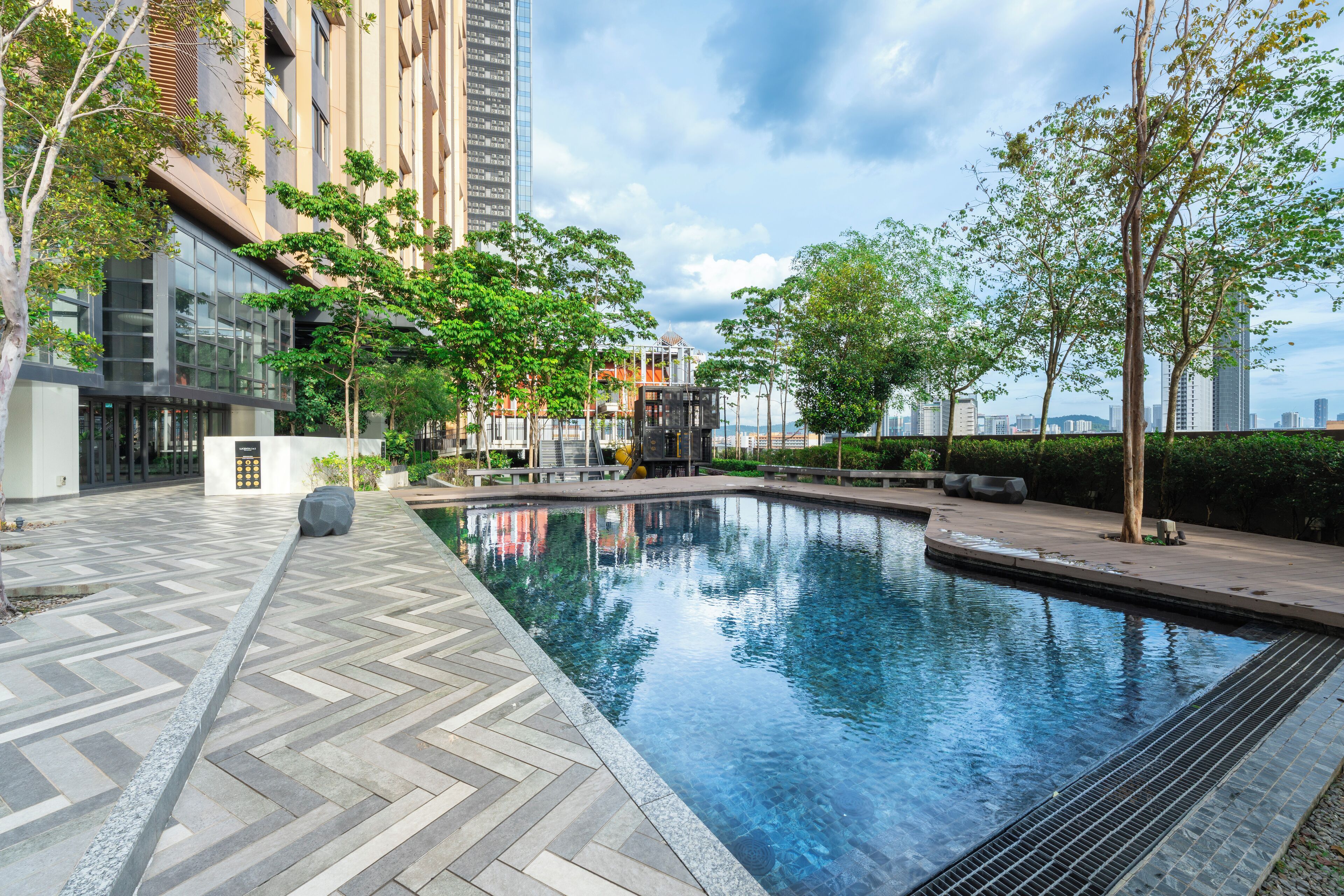 Una piscina exterior moderna con un diseño de mosaicos en patrón de espiga, rodeada de árboles y un edificio con fachada de vidrio.