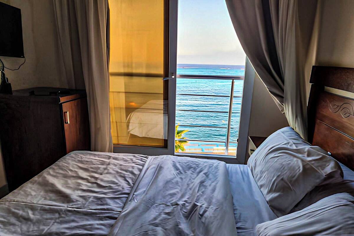 Habitación de hotel con una cama, cabecera de madera, mesita de noche y vista al océano a través de una puerta corrediza.