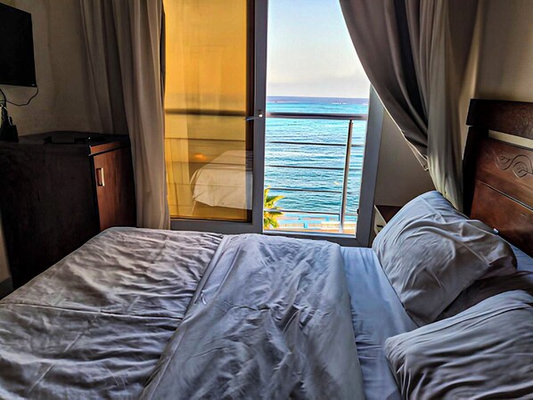 A hotel room with a bed, a wooden headboard, a nightstand, and a view of the ocean through a sliding door. - Luxury Apartment 3BR & Full Sea View (Marsa Matrouh)