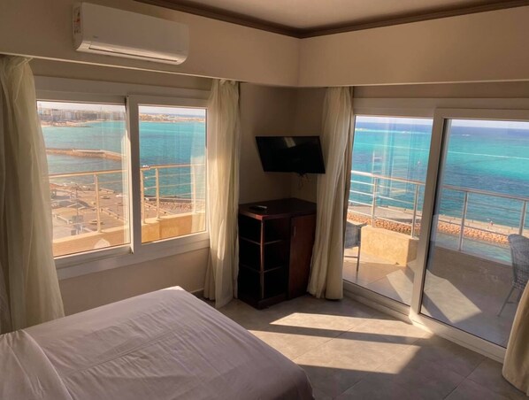 A hotel room with a bed, a TV, and large windows offering a view of the sea and a beach. - Luxury Apartment 3BR & Full Sea View (Marsa Matrouh)
