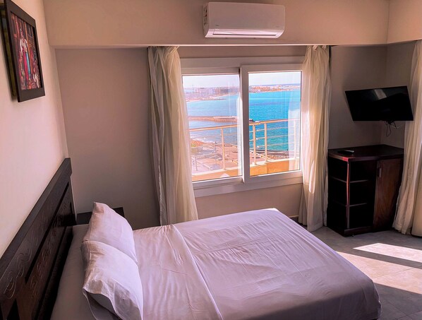 A hotel room with a bed, a TV, a desk, and a window with a view of the sea. - Luxury Apartment 3BR & Full Sea View (Marsa Matrouh)