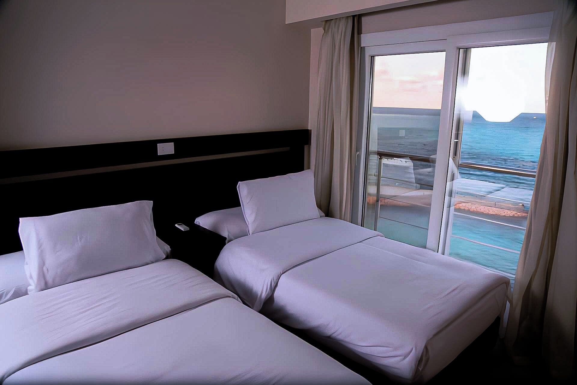 A hotel room with two beds, a large window with a view of the sea, and a balcony.
