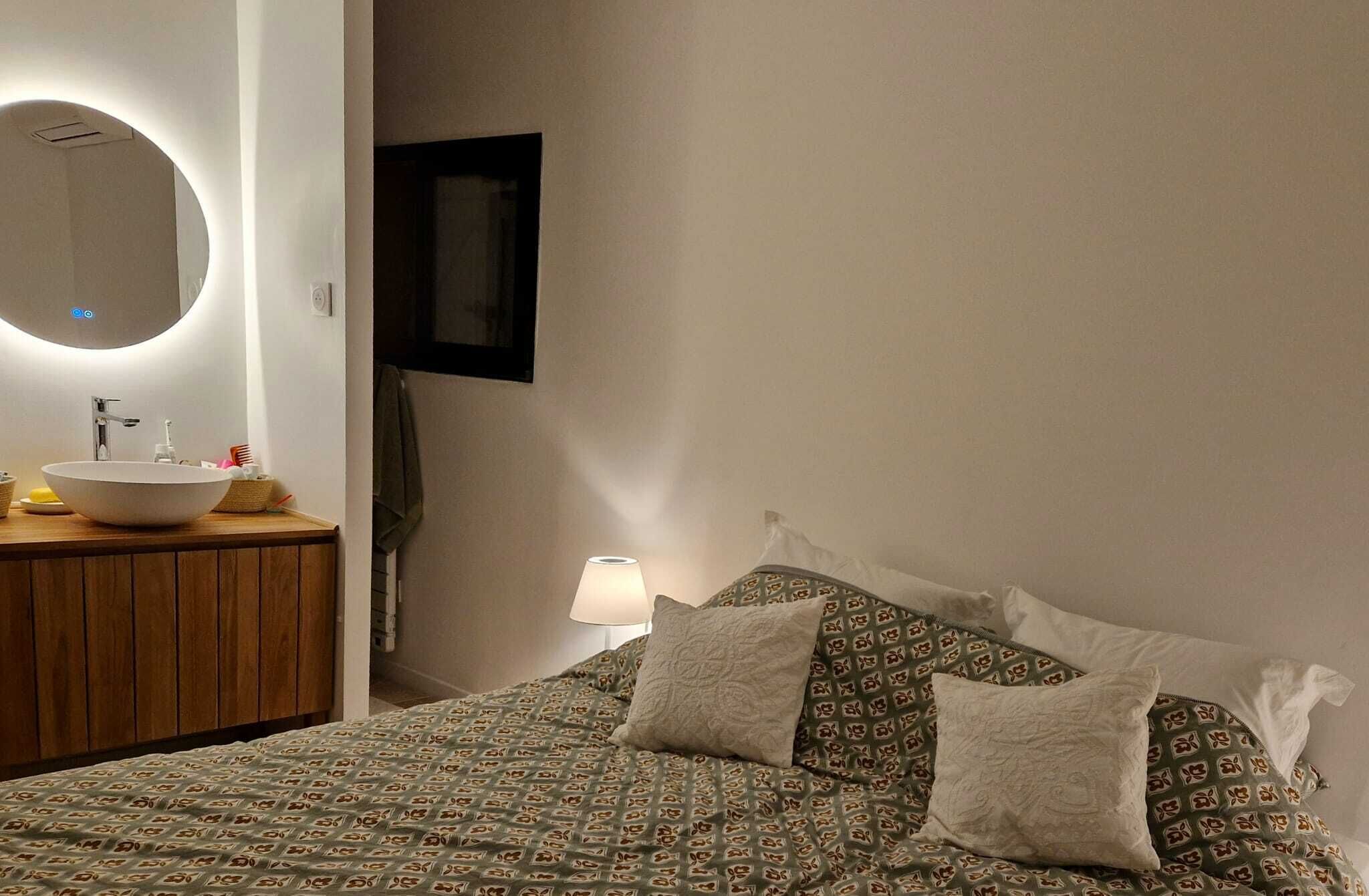 A bedroom with a round mirror, a bedside table with a sink, a bed with patterned bedding, and a wall-mounted TV.