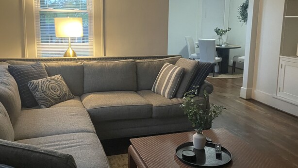 A living room with a sectional sofa, a coffee table, and a lamp. There is a dining area with a table and chairs in the background. - Welcome to Augusta. This beautiful and home has lots of Southern Charm. (Augusta)